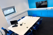 Meeting room with tables, chairs, a small screen and a larger screen on the wall